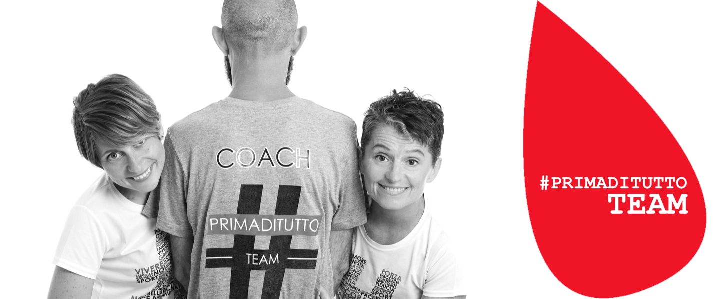 Tre persone in una foto in bianco e nero: al centro, una persona di spalle indossa una maglietta con la scritta 'COACH PRIMADITUTTO TEAM'. Ai lati, due donne sorridono guardando la fotocamera.